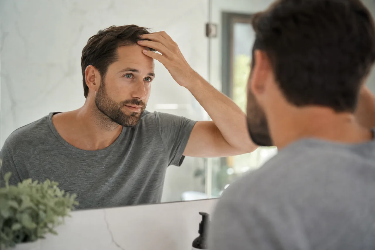 A man checking his hairline in a bright bathroom mirror